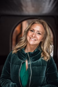 Cathrine Fossum stående foran en buet bakgrunn, smilende, med grønn velourjakke og blondt hår med store bølger.