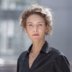 Profilbilde Ingrid Hjort, krøllete brunt hår, rød leppestift, mørk skjorte, briller