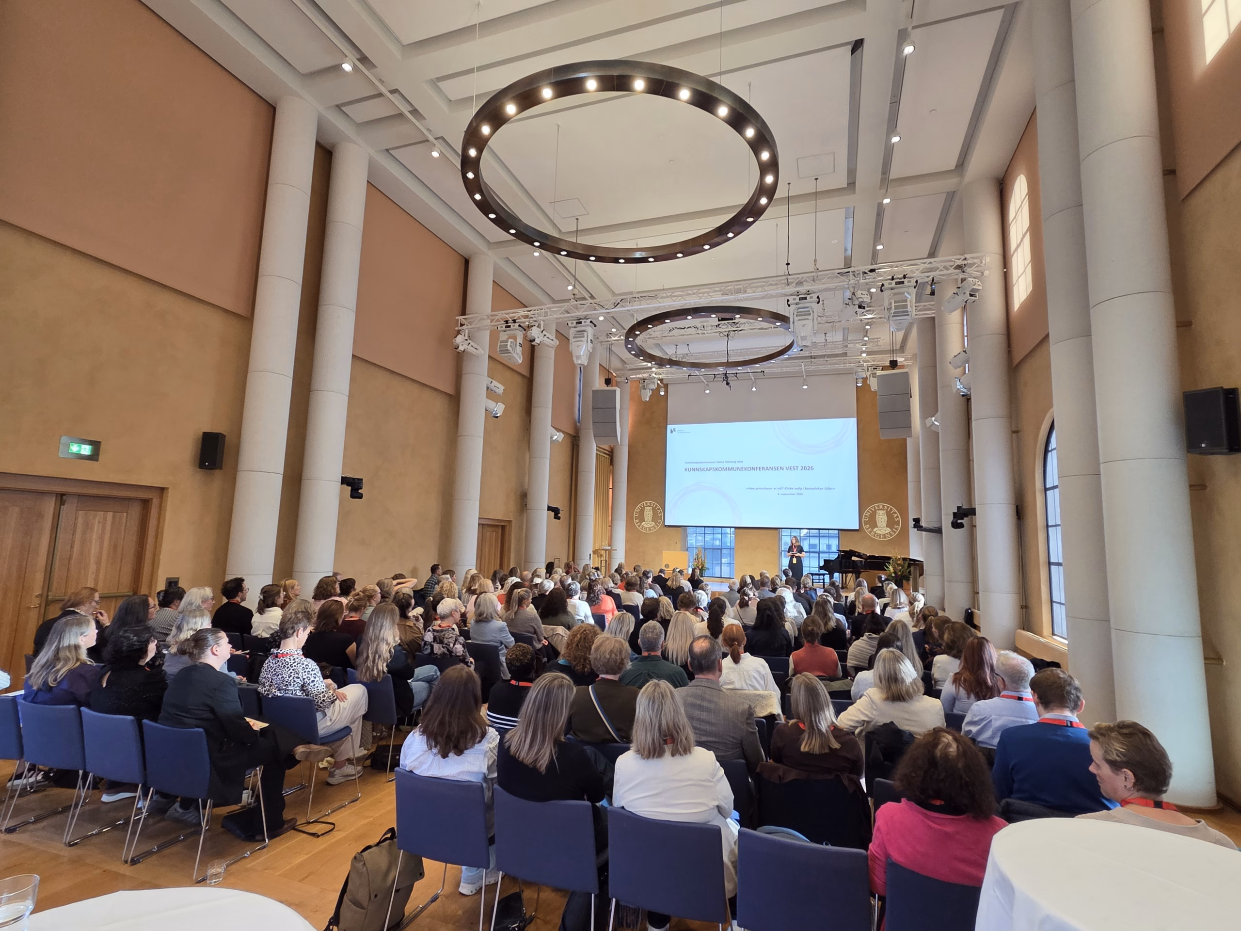 Fullsatt universitetsaula, bilde fotografert fra bak i salen, med fokus på tak og skjerm foran i salen
