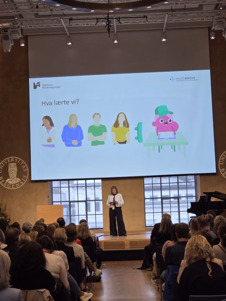 Bilde av Kristin H. Bruvik på scenen under sitt innlegg, tatt bak i lokalet slik at man også ser noe av publikum.