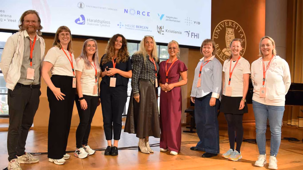 Bilde av 9 personer på scenen i universitetsaulaen, arrangører sammen med konferansier Cathrine Fossum